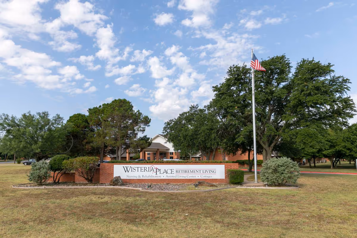 Entrance sign and front lawn of Wisteria Place assisted living with a flagpole, trees, and the building in the background.
