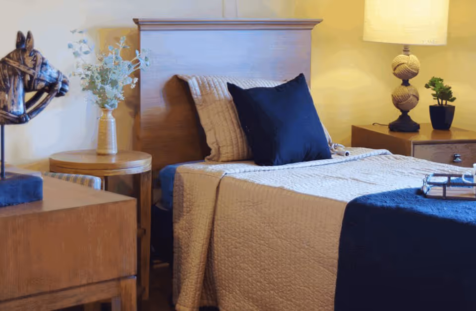 Neatly made single bed with a beige quilt, navy pillows and throw, wooden headboard and bedside tables with a lamp and decorative items.