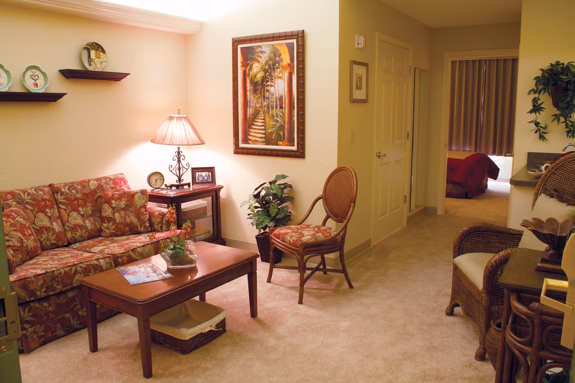 A cozy living room area in a senior living facility with a floral patterned sofa, a wooden coffee table with a basket underneath, a side table with a lamp, clock, and framed photo, a wooden chair with a matching floral cushion, a framed painting on the wall, and a view into a bedroom with a red bedspread.