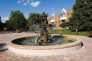 A large decorative fountain with multiple tiers and water spouts in a circular basin, situated in a paved courtyard area with green trees and a multi-story brick building in the background under a blue sky with some clouds.