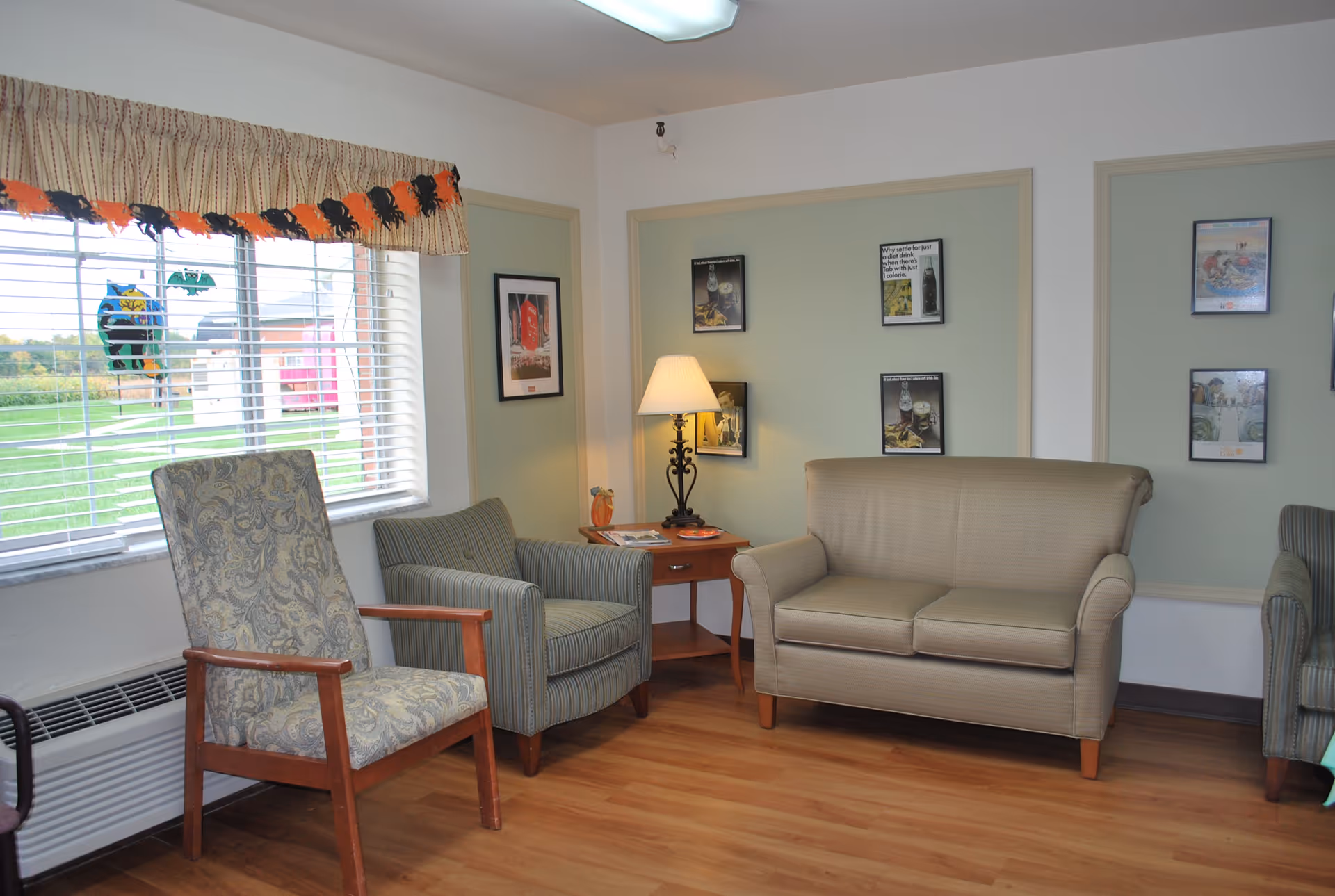 A cozy sitting area in a nursing and rehabilitation facility with a patterned armchair, a striped armchair, and a beige loveseat arranged around a wooden side table with a lamp. The walls have framed pictures, and a window with blinds and a decorative valance overlooks a green outdoor area.