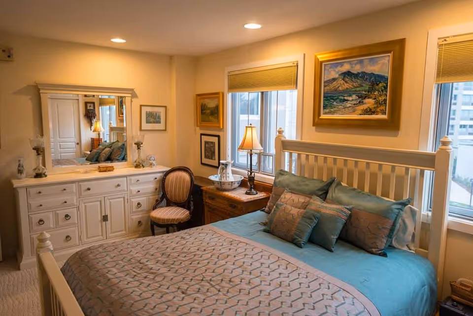A cozy bedroom with a white wooden bed frame, teal and beige bedding, and multiple pillows. There is a wooden nightstand with a lamp and decorative items next to the bed. A white dresser with a large mirror above it and a cushioned chair are also visible. The room has two windows with beige blinds and framed paintings on the walls.