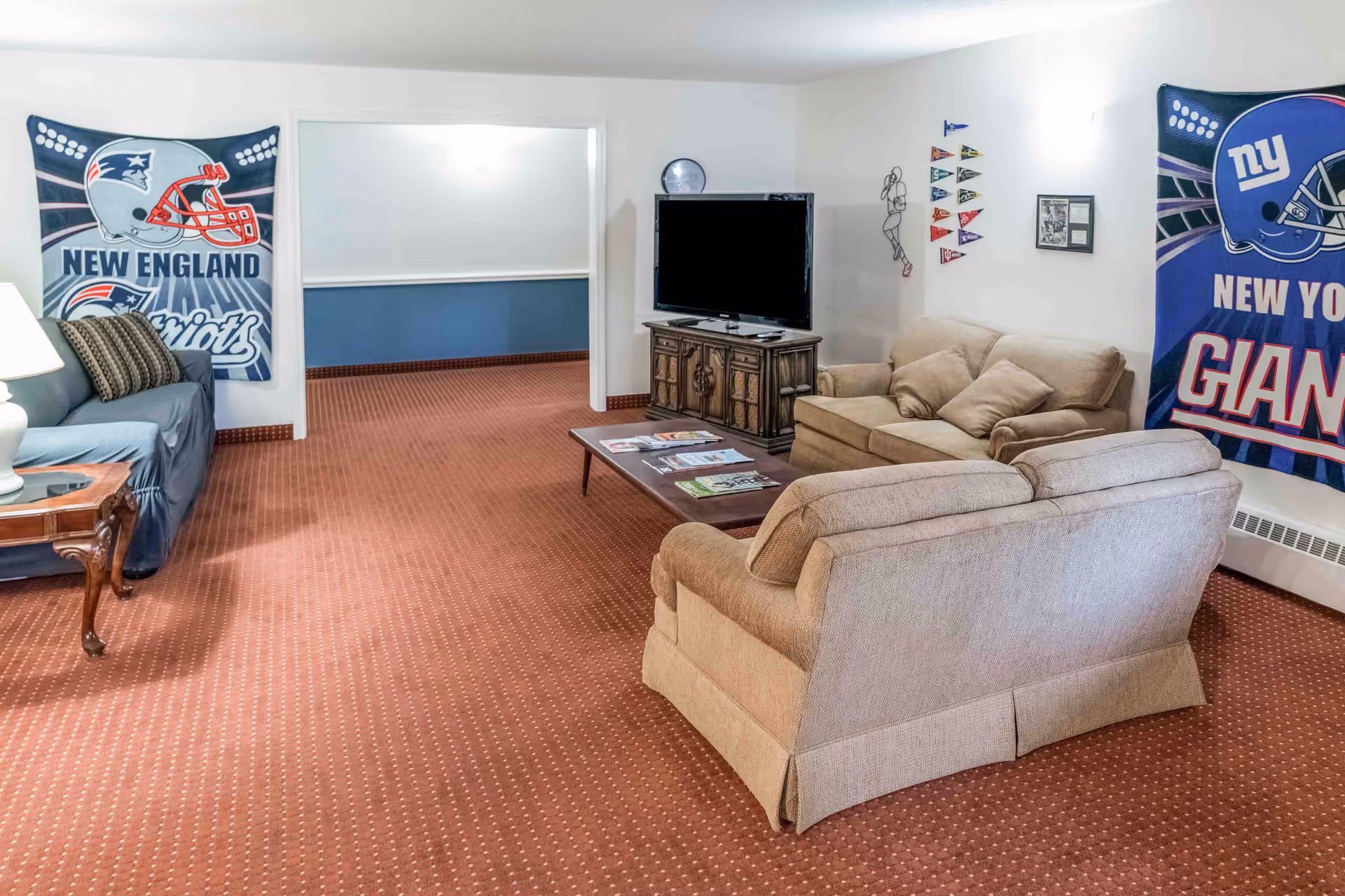 Carpeted lounge with sofas, a TV on a wooden cabinet, a coffee table with magazines, and football team banners on the walls.