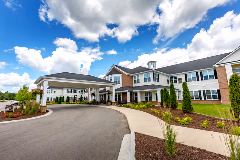 Exterior view of StoryPoint Rockford senior living facility showing a large two-story building with a covered entrance driveway, landscaped garden beds with shrubs and flowers, and a partly cloudy blue sky.