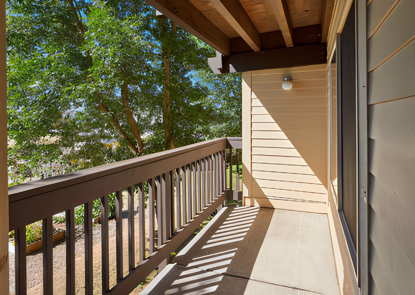 A sunlit balcony with wooden railings and beige siding on the right side, overlooking green trees and a garden area below.