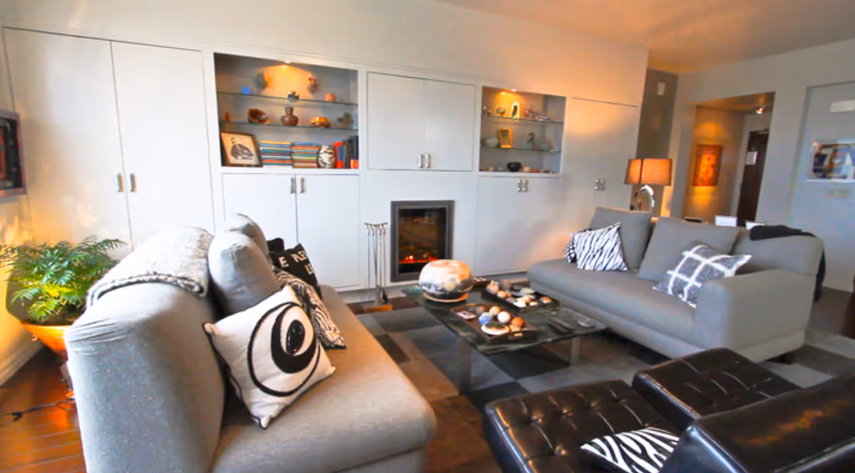 A modern living room with two gray sofas adorned with patterned pillows, a black leather ottoman, a low coffee table with decorative items, built-in white cabinets with shelves displaying books and ornaments, a small fireplace, a floor lamp, and a potted plant in the corner.