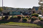 Exterior view of Cedar Hill Healthcare & Rehab Center showing the main entrance with a covered drop-off area, a landscaped garden with bushes and grass in front, and a sign displaying the facility's name.