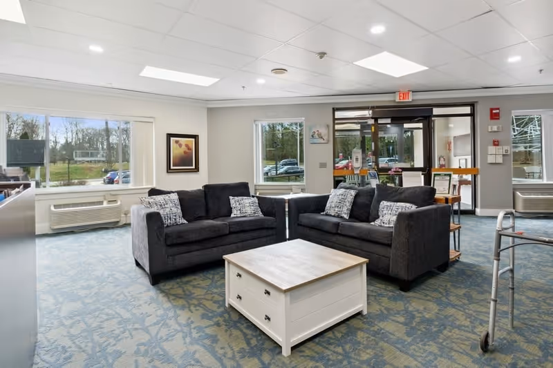 A bright and spacious senior living facility common area with two dark gray sofas facing each other around a white wooden coffee table. The room has large windows letting in natural light, patterned blue carpet, and a glass entrance door in the background. A walker is visible on the right side of the image.