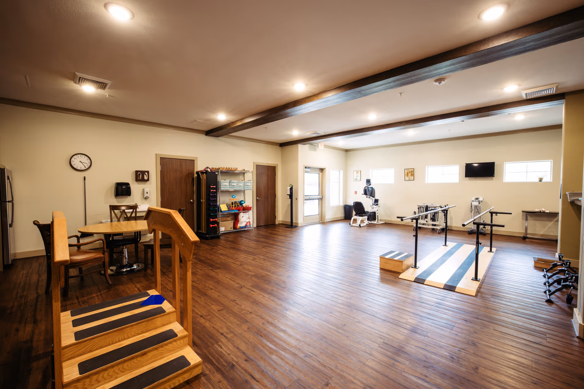 A spacious rehabilitation room with wooden flooring and beige walls. The room features a small set of wooden stairs with handrails, parallel bars for walking exercises, a chair with exercise equipment, a wall-mounted TV, and a table with chairs in the corner. Shelves with various exercise tools and supplies are also visible.