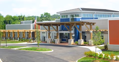Exterior view of Stonecroft Health Campus showing a modern building with a covered entrance supported by wooden beams, surrounded by landscaped greenery and a paved driveway.