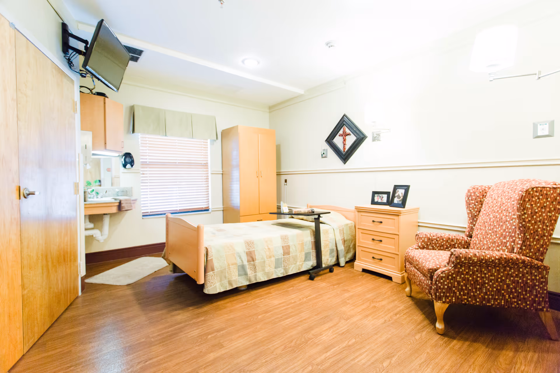 A senior living facility bedroom with a single bed covered with a patchwork quilt, a wooden nightstand with framed photos, a patterned armchair, a wall-mounted TV, a window with blinds and a valance, a wooden wardrobe, and a small sink area with a mirror.