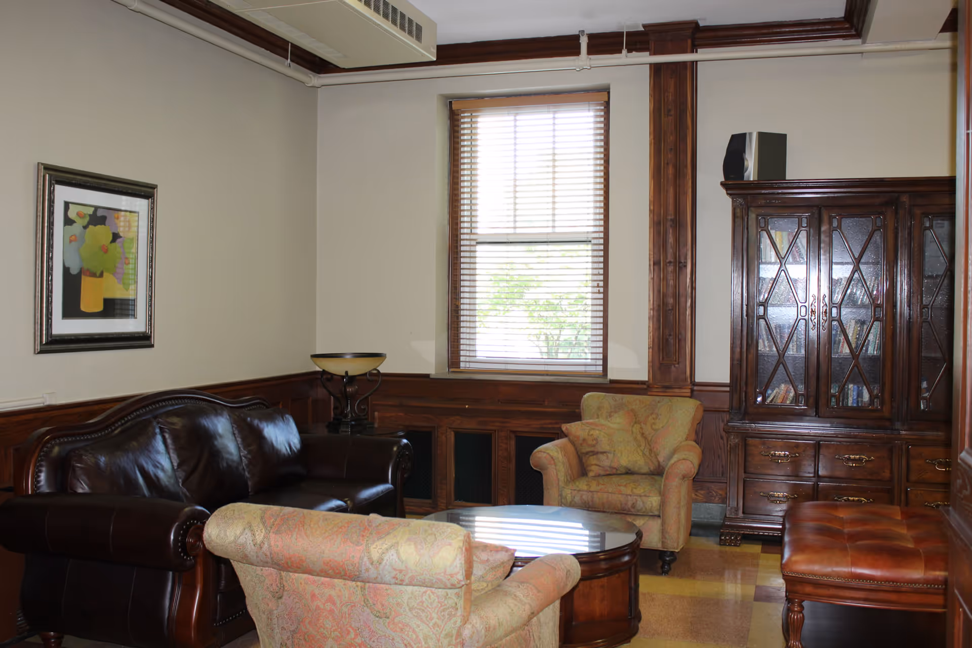 A cozy living room with a dark brown leather sofa, two patterned armchairs, a round wooden coffee table, a wooden cabinet with glass doors, a window with blinds, a framed floral painting on the wall, and a cushioned bench.