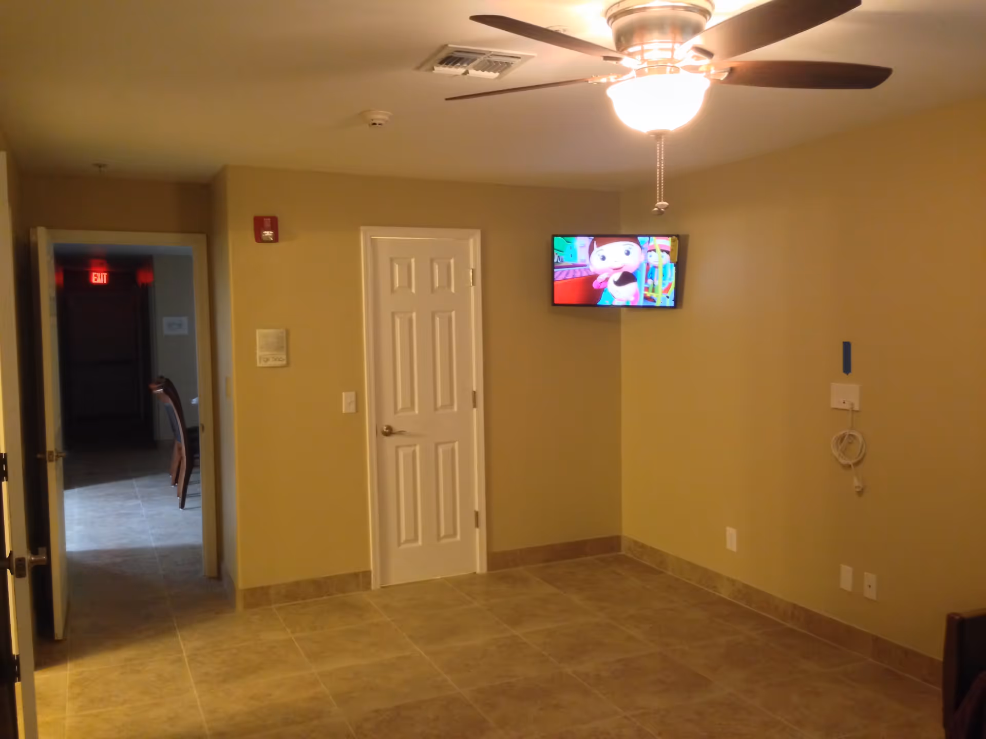 An empty room with beige walls and tiled floor, a ceiling fan with light, a white door, and a wall-mounted TV displaying a cartoon. There is an open doorway leading to a hallway with an exit sign visible.