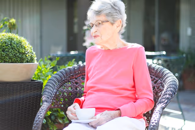 An elderly woman with short gray hair and glasses is sitting outdoors on a wicker chair, holding a white teacup and saucer. She is wearing a bright pink long-sleeve shirt and white pants. There are green plants and outdoor furniture in the background.
