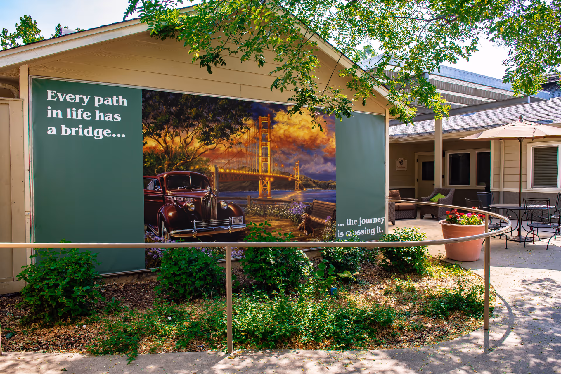 Exterior courtyard of a memory care facility featuring a large mural reading "Every path in life has a bridge..." with a vintage car, seating area, umbrella and landscaping.