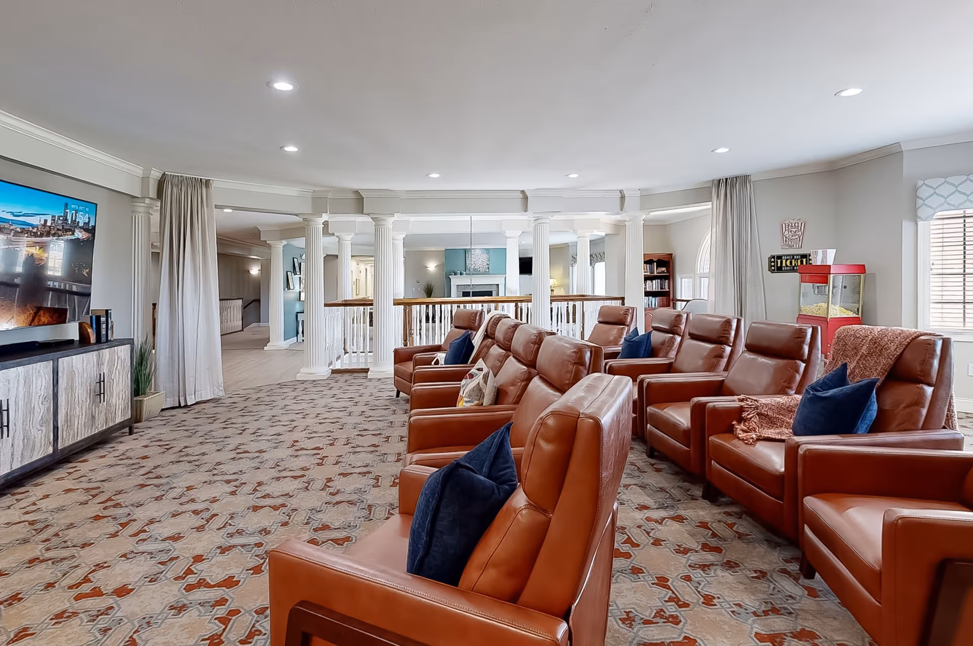 A spacious living room with multiple brown leather recliner chairs arranged in rows facing a large flat-screen TV mounted on a light-colored wall. The room features a patterned carpet, white columns, large windows with curtains, a popcorn machine, and a bookshelf in the background.