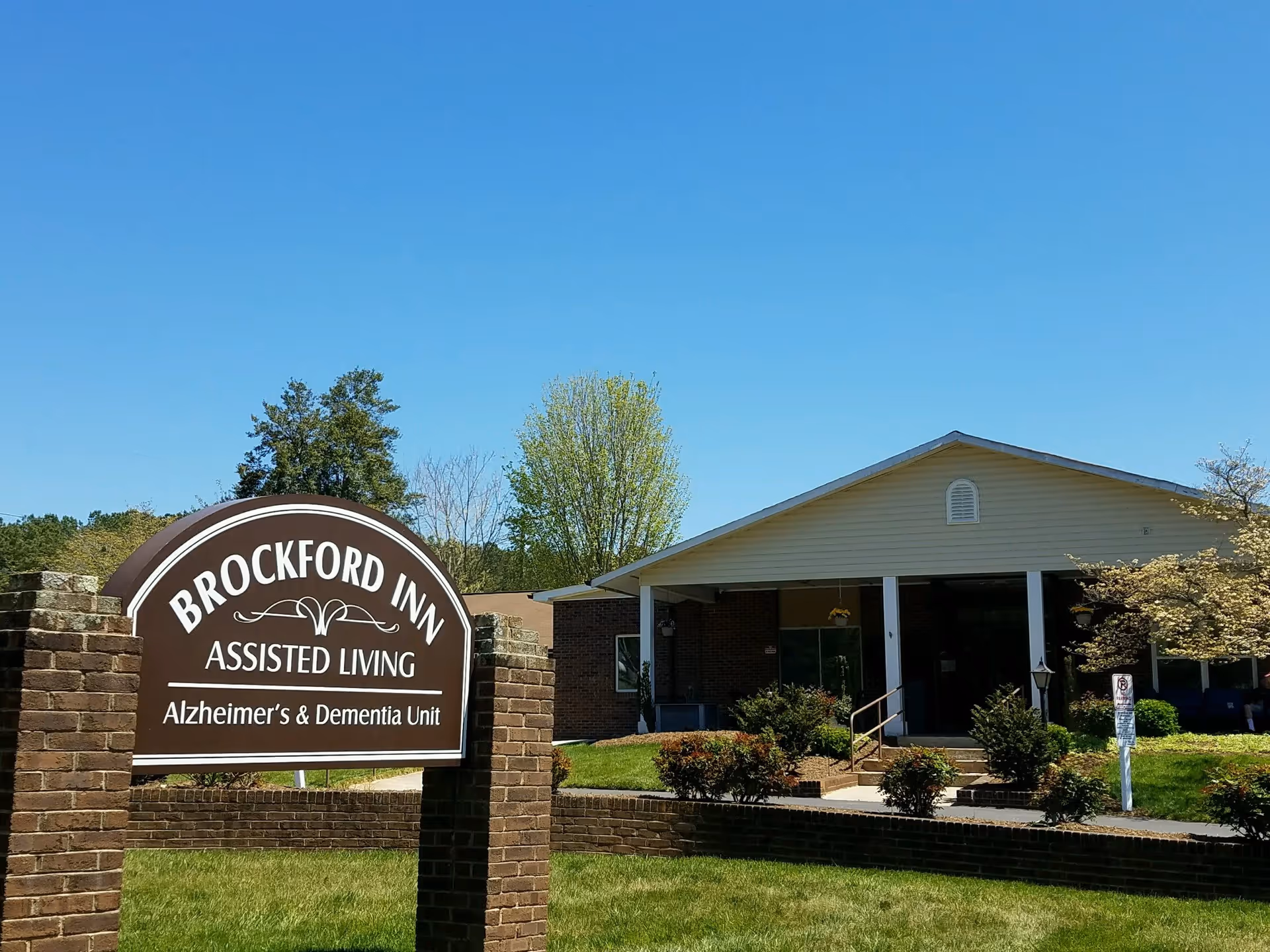Exterior view of Brockford Inn Assisted Living facility with a sign in the foreground that reads 'Brockford Inn Assisted Living Alzheimer's & Dementia Unit'. The building is single-story with a covered entrance, surrounded by green grass, bushes, and trees under a clear blue sky.