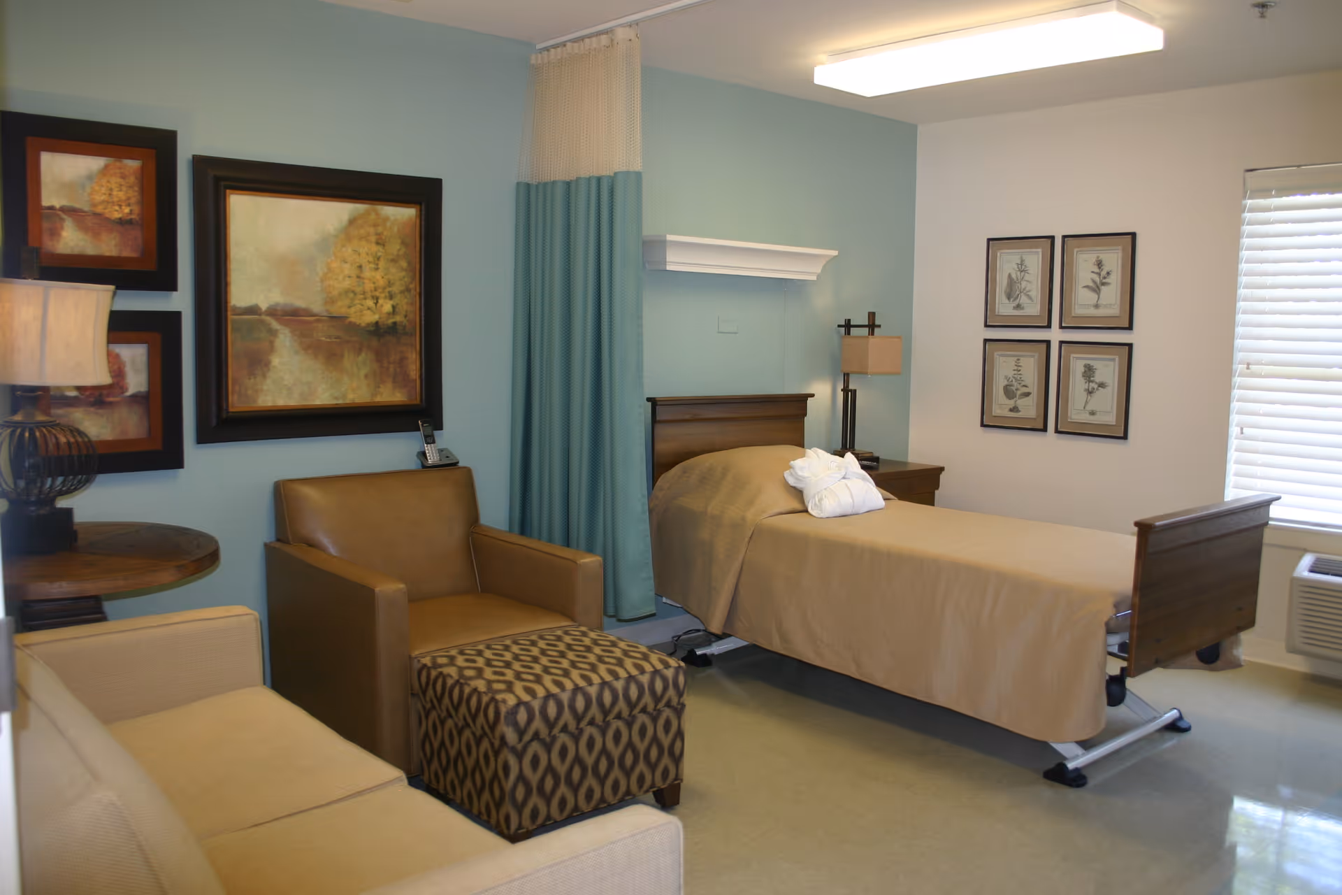 A senior living facility bedroom with a single bed covered in a beige blanket, a folded white robe on top, a brown armchair with a patterned ottoman, a beige sofa, a round wooden side table with a lamp, and framed artwork on the walls. There is a window with blinds and an air conditioning unit below it.