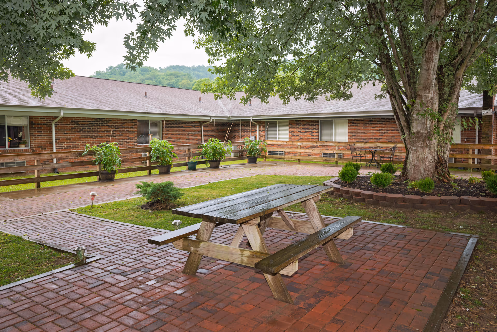 Outdoor brick courtyard with a wooden picnic table under a large tree beside a single-story brick building.