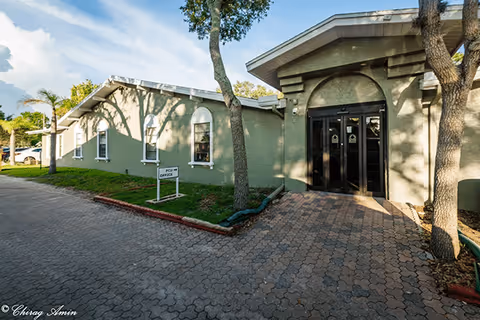 Entrance of a single-story green building with a paved walkway, trees, and a small lawn sign.