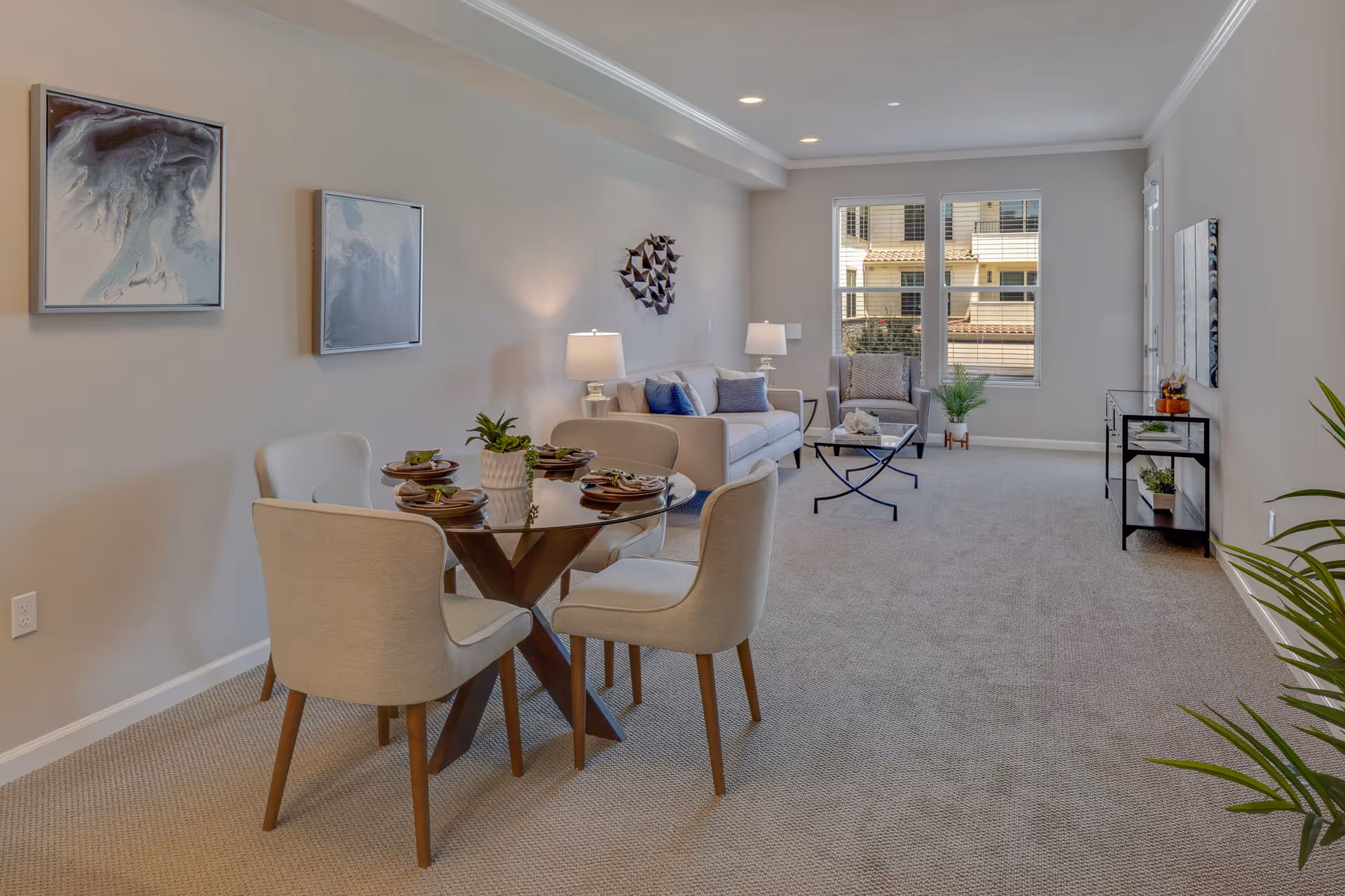 A spacious, well-lit living room and dining area with beige carpet and light-colored walls. The dining area features a round glass table with four upholstered chairs and place settings. The living room has a sofa, armchair, coffee table, side tables with lamps, and decorative wall art. Large windows allow natural light to enter the room.