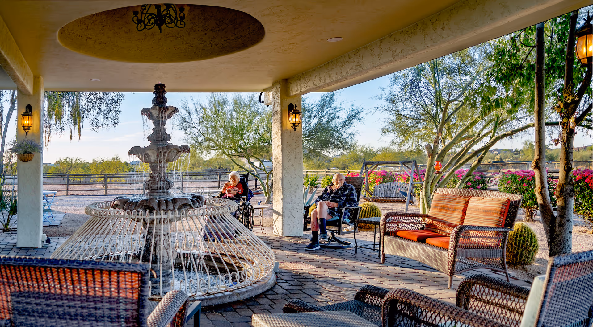 Outdoor covered patio area with a large decorative water fountain in the center. Two elderly individuals are seated nearby, one in a wheelchair and the other in a chair, surrounded by wicker furniture with orange cushions. The area is landscaped with desert plants, trees, and colorful flowers under a clear sky.