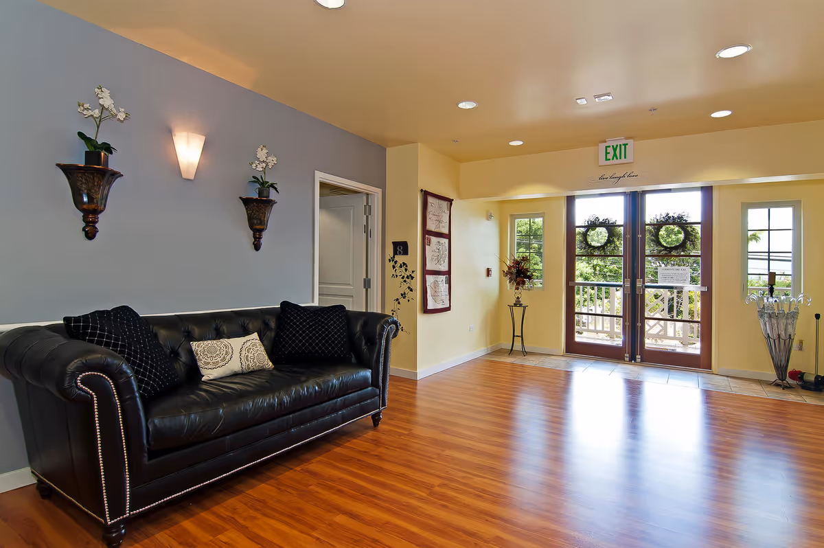 A bright and welcoming senior living facility common area with a black leather couch adorned with three decorative pillows. The walls are painted in soft yellow and gray tones, with two wall-mounted planters holding white flowers and a wall sconce light above the couch. The room features wooden flooring and a double glass door exit with wreath decorations, flanked by windows letting in natural light. A small table with a flower arrangement and an umbrella stand are near the entrance.
