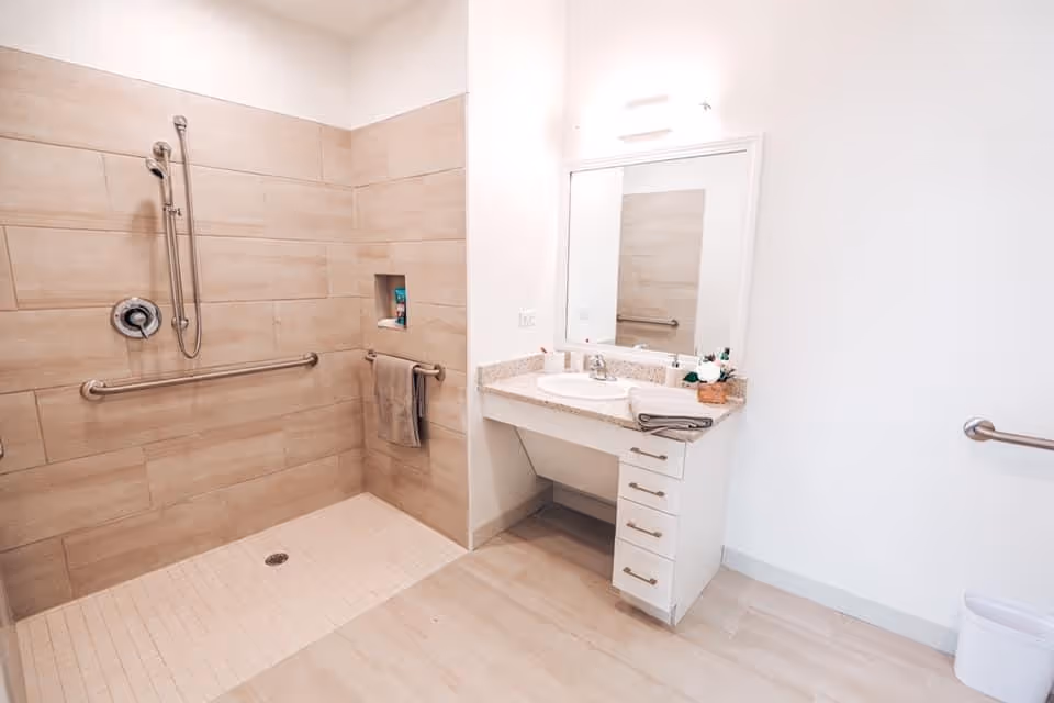 A clean, accessible bathroom featuring a walk-in shower with beige tiles, a handheld showerhead, and grab bars. There is a towel hanging on a bar inside the shower area. To the right, there is a white vanity with a sink, a large mirror, and drawers underneath. The bathroom has light-colored flooring and walls, with additional grab bars on the walls for support.
