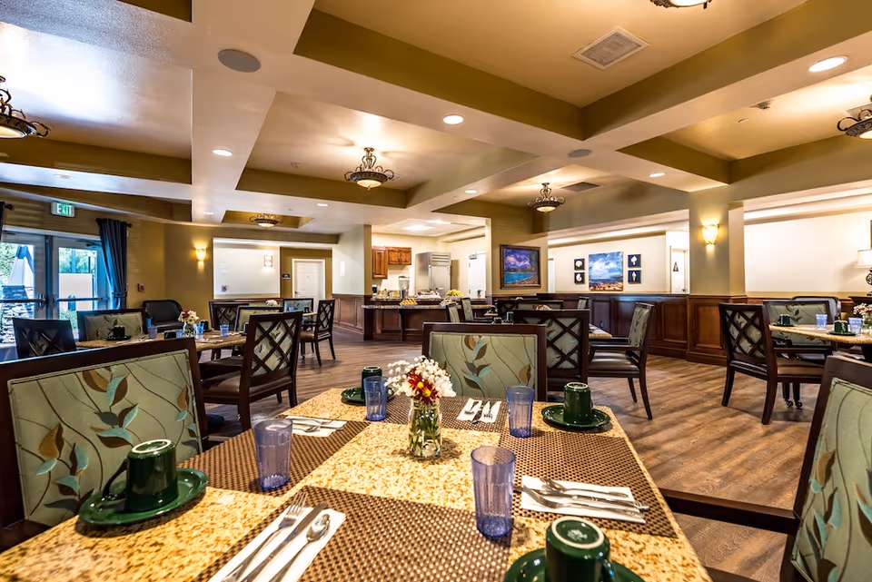 Communal dining room with tables set with plates, cups, and utensils, surrounded by upholstered chairs and warm lighting.