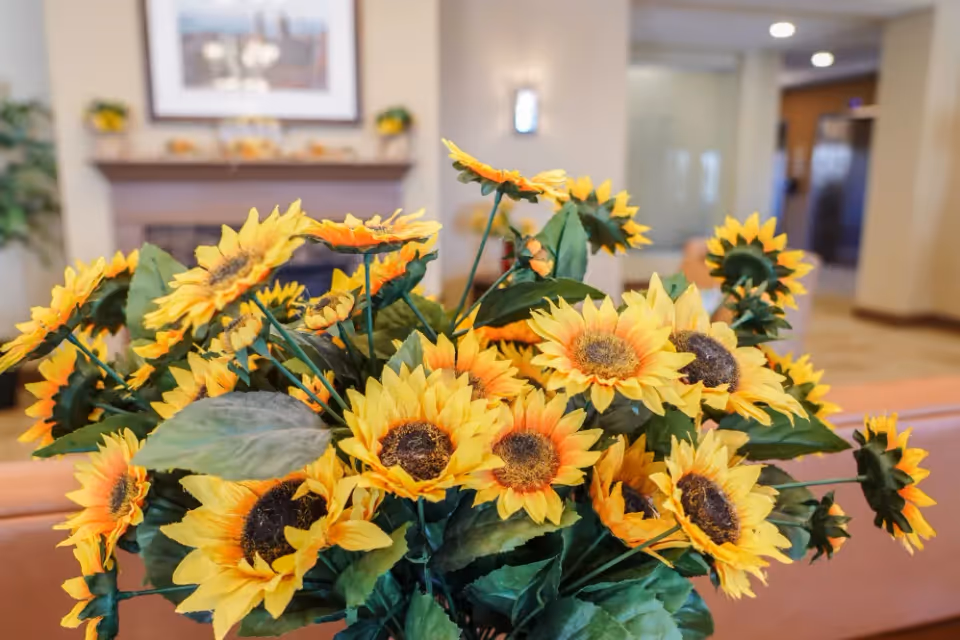 A close-up view of a bouquet of bright yellow sunflowers with green leaves in an indoor setting. In the background, there is a blurred fireplace with a framed picture above it and some plants on either side, suggesting a cozy living room or common area.