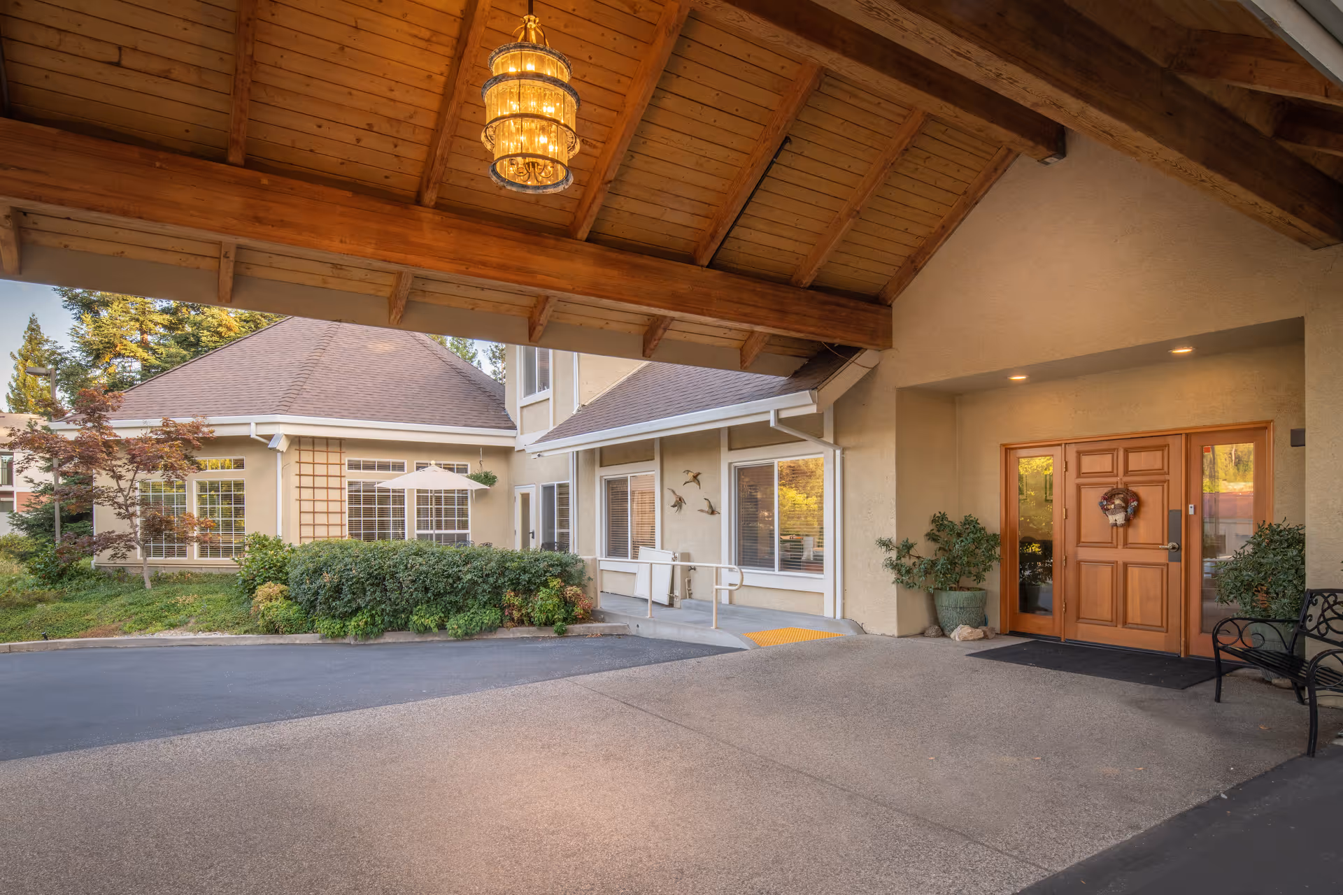 Covered entrance area of Brunswick Village Assisted Living with a wooden ceiling and hanging light fixture. The entrance features double wooden doors decorated with a wreath, flanked by potted plants and a bench. The building exterior is beige with large windows and surrounding greenery including bushes and trees.