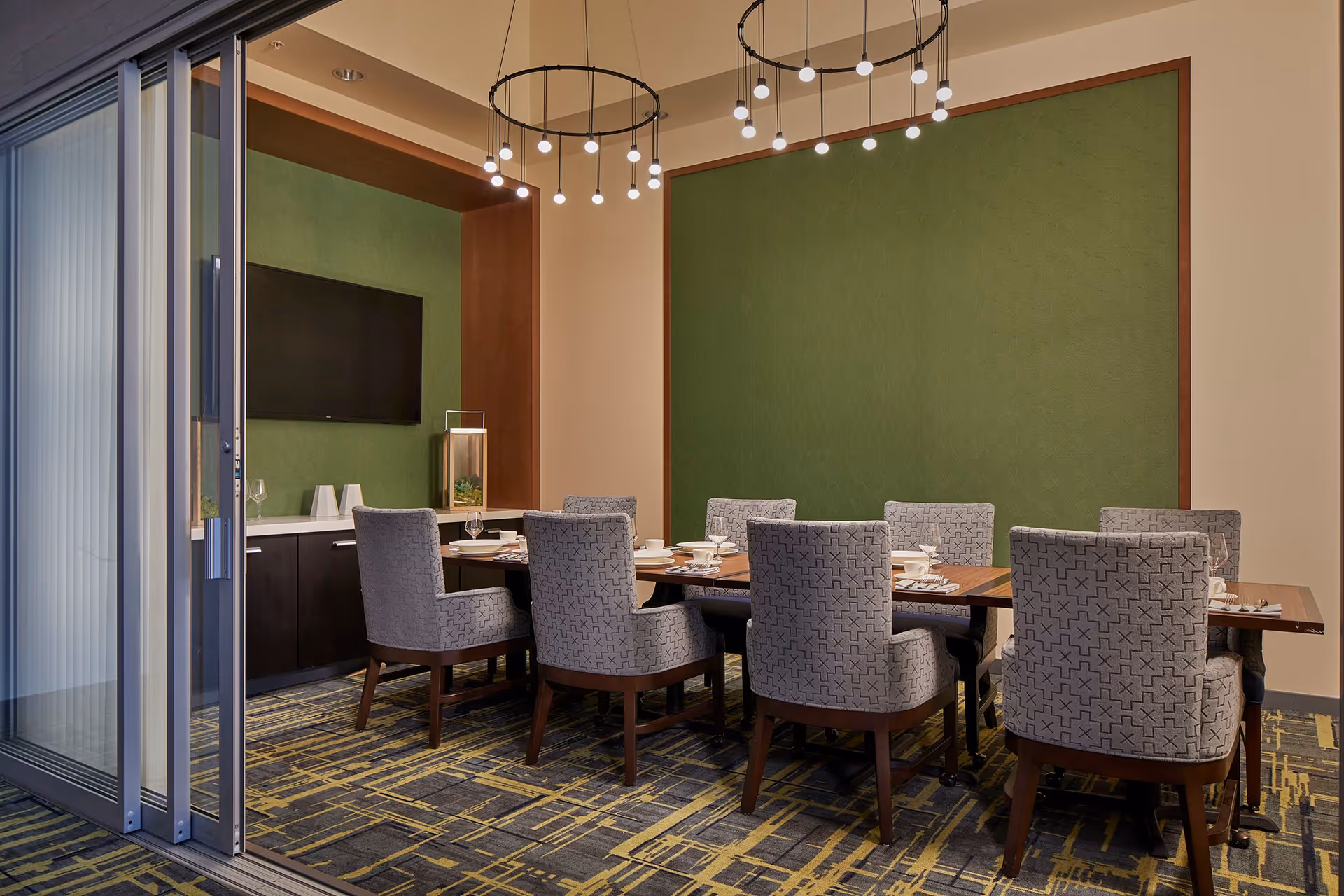 A modern dining room with a rectangular wooden table surrounded by eight upholstered chairs with patterned fabric. The room features a green accent wall, a flat-screen TV mounted on another green wall, a sideboard with decorative items, and two circular pendant light fixtures hanging from the ceiling. The floor has a patterned carpet, and there is a sliding glass door partially open on the left side.
