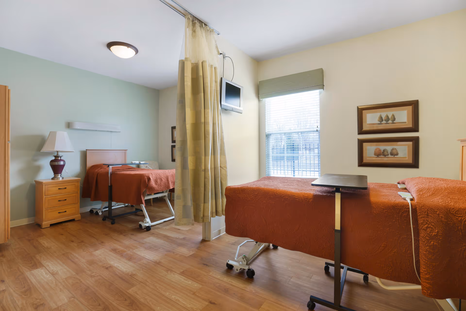 A senior living facility bedroom with two hospital-style beds covered in orange bedspreads. The room has wooden flooring, a bedside table with a lamp, a wall-mounted TV, a window with blinds and a green valance, and two framed pictures on the wall. A privacy curtain separates the two beds.
