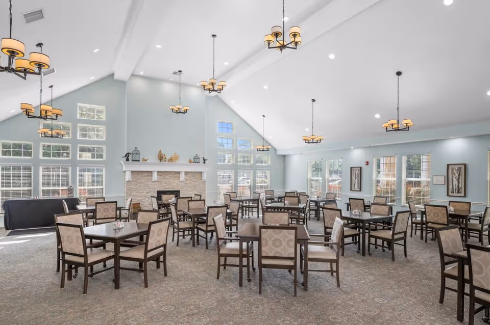 Spacious senior living dining room with multiple tables and chairs, chandeliers, large windows, and a fireplace.