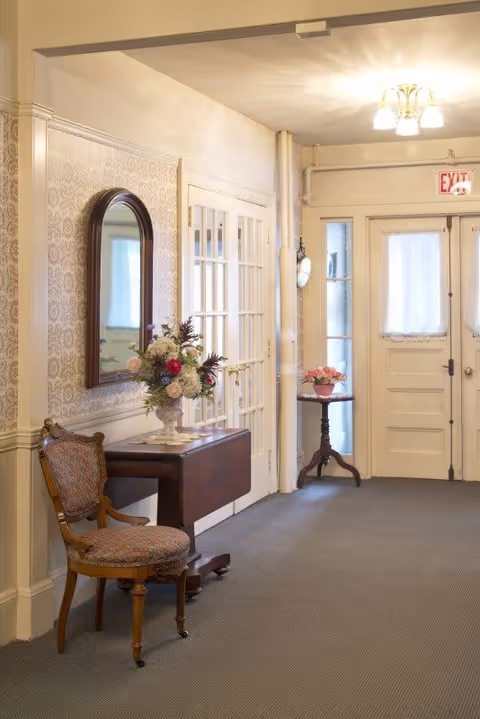 A hallway with beige walls and carpeted floor featuring a wooden chair with patterned upholstery, a small wooden table with a floral arrangement, a wall-mounted mirror with a wooden frame, and a small round table with a pink flower arrangement near a white door with frosted glass panels and an exit sign above it.