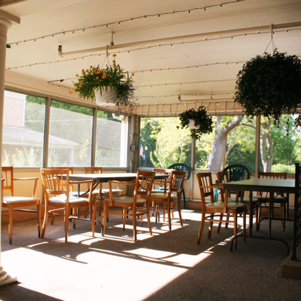 A sunlit enclosed patio or sunroom with several wooden chairs and tables arranged for seating. Hanging flower pots with green plants and flowers are suspended from the ceiling, and large windows allow natural light to fill the space, showing greenery outside.