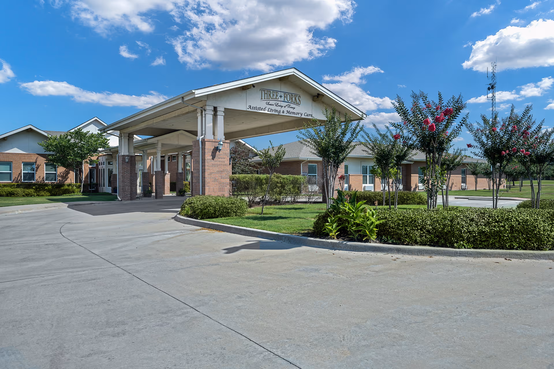 Front entrance and covered porte-cochere of a single-story senior living facility with manicured landscaping under a blue sky.