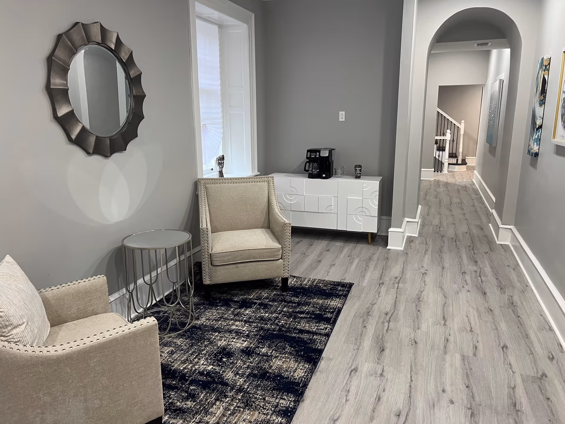 A modern interior seating area with two beige armchairs, a small round glass-top side table, a decorative round mirror on the wall, a white cabinet with a coffee maker and cups, and a hallway with light wood flooring and artwork on the walls.
