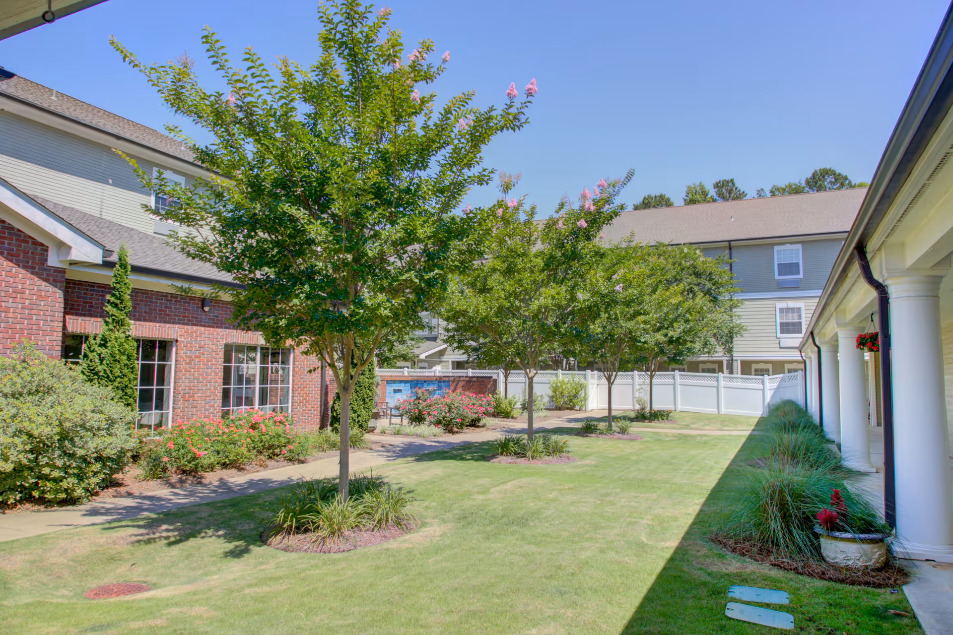 A sunny outdoor courtyard area at Truewood by Merrill, Riverchase featuring green grass, several small trees with pink flowers, shrubs, and a paved walkway. The courtyard is surrounded by buildings with brick and siding exteriors, white fencing, and columns on the right side.
