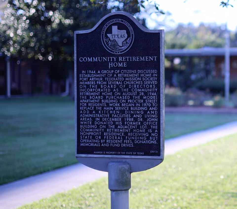 A historical marker sign for Community Retirement Home in Texas, detailing its establishment in 1944, development history, and nonprofit status, set outdoors with grass and a building blurred in the background.