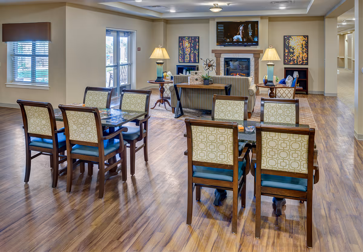 A senior living facility common area with two dining tables and eight chairs in the foreground, a seating area with a sofa and armchairs around a fireplace with a TV mounted above it in the background, and two table lamps on side tables. The room has wood flooring, beige walls, and windows letting in natural light.