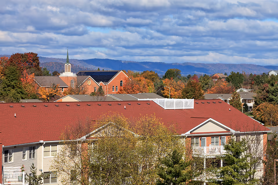 View of a residential building with a red roof surrounded by trees with autumn foliage, set against a backdrop of hills and a partly cloudy sky. In the distance, there is a church with a tall steeple and solar panels on its roof.