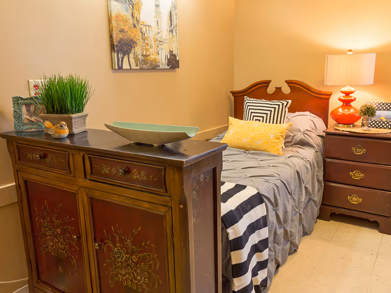 A cozy single bedroom with a wooden headboard bed, striped throw and decorative pillows, a nightstand with a lamp, and a painted wooden cabinet.
