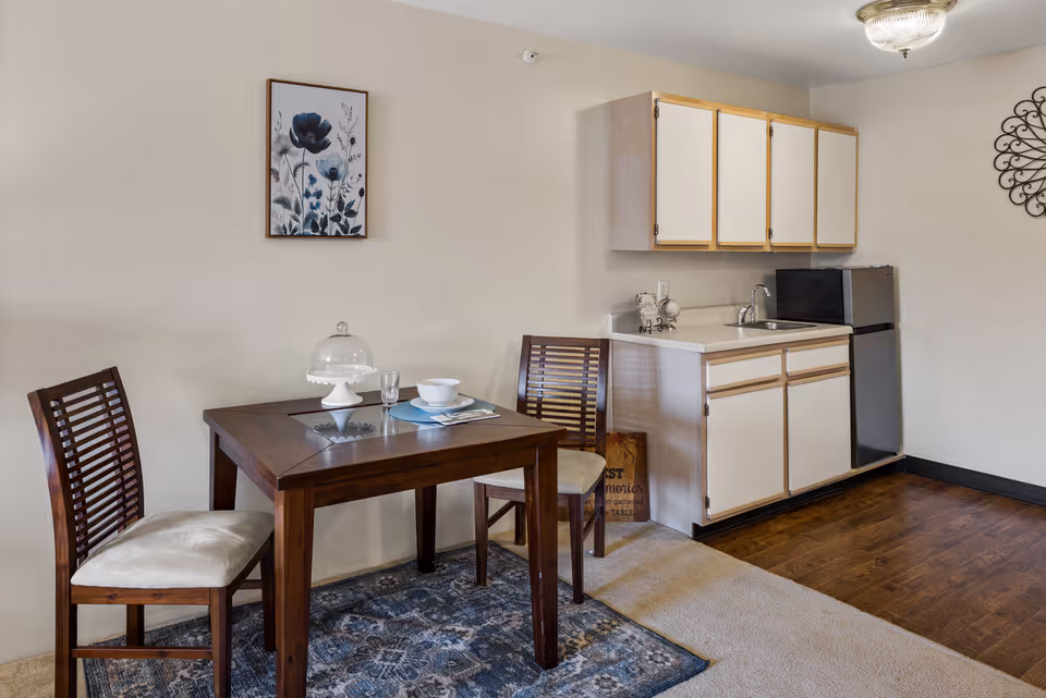 A small dining area with a wooden table set for one, featuring a glass, bowl, and plate on a blue placemat. Two wooden chairs with cushioned seats are placed around the table. Behind the dining area is a compact kitchen with white cabinets, a sink, and a small refrigerator. The floor transitions from carpet near the dining area to wood in the kitchen. A framed floral artwork hangs on the wall above the table, and a decorative metal wall piece is visible on the right side.