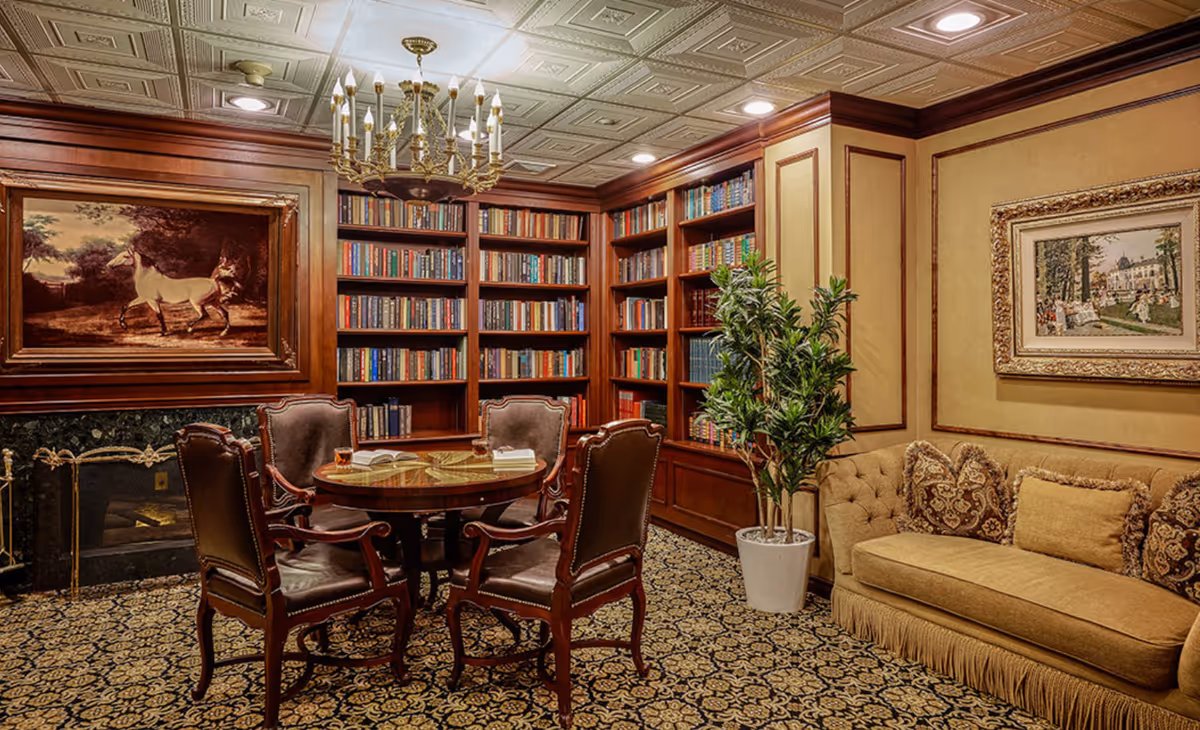 A cozy library room with wooden bookshelves filled with books, a round wooden table surrounded by four upholstered chairs, a beige sofa with decorative pillows, a potted plant, and framed paintings on the walls. The ceiling has a decorative pattern with recessed lighting and a chandelier.