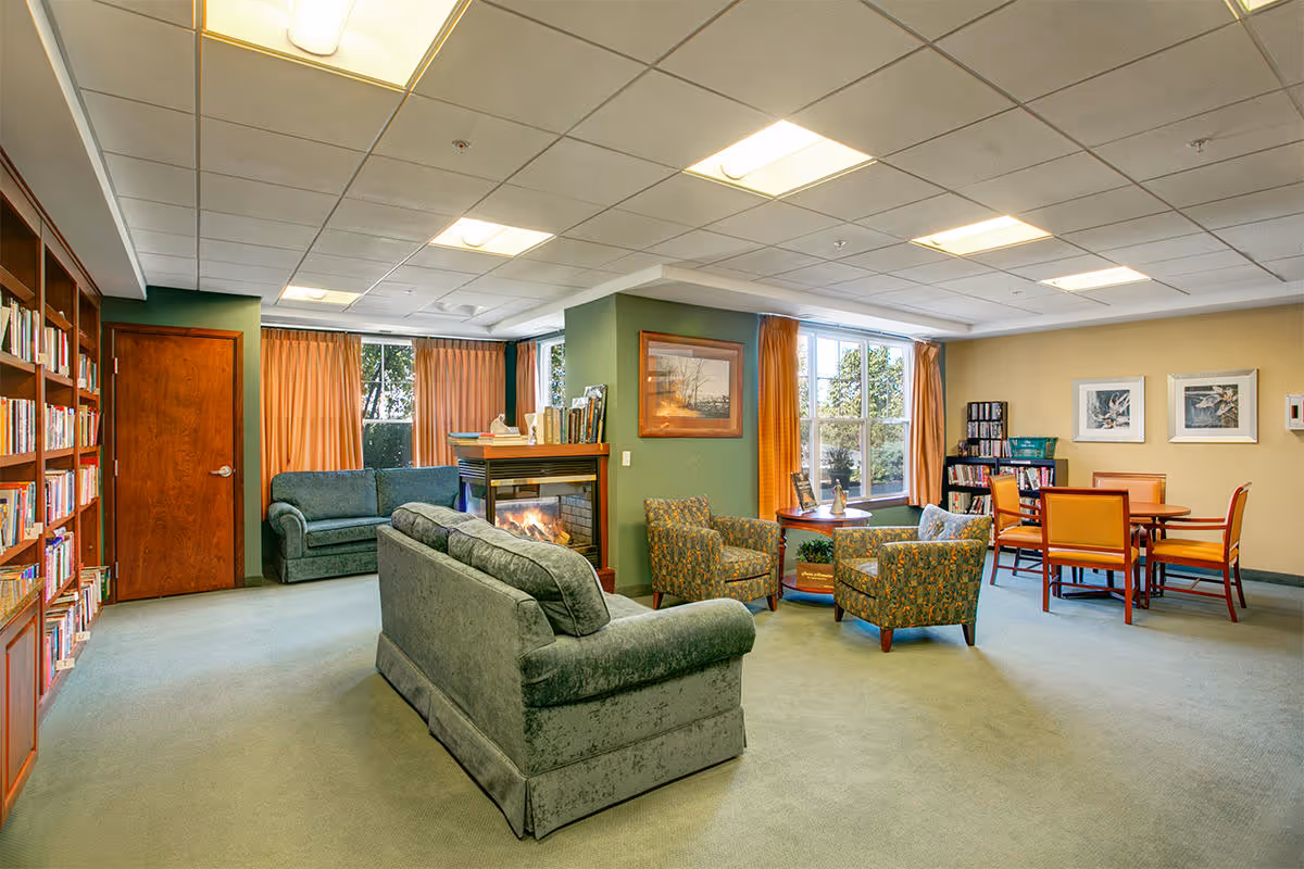 A cozy living room area in a senior living facility featuring green upholstered sofas and armchairs arranged around a fireplace. There are large windows with orange curtains letting in natural light, a wooden bookshelf filled with books, and a dining table with chairs in the background. The walls are painted in green and beige tones, and framed artwork is displayed on the walls.