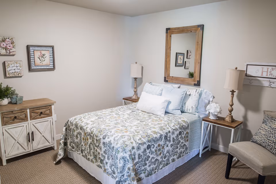 A cozy bedroom with a bed covered in a floral patterned quilt and multiple pillows. On either side of the bed are wooden nightstands with matching lamps. A large wooden framed mirror hangs above the bed. To the left, there is a wooden dresser with drawers and decorative framed floral artwork on the wall above it. A cushioned chair with a patterned pillow is positioned on the right side of the room.