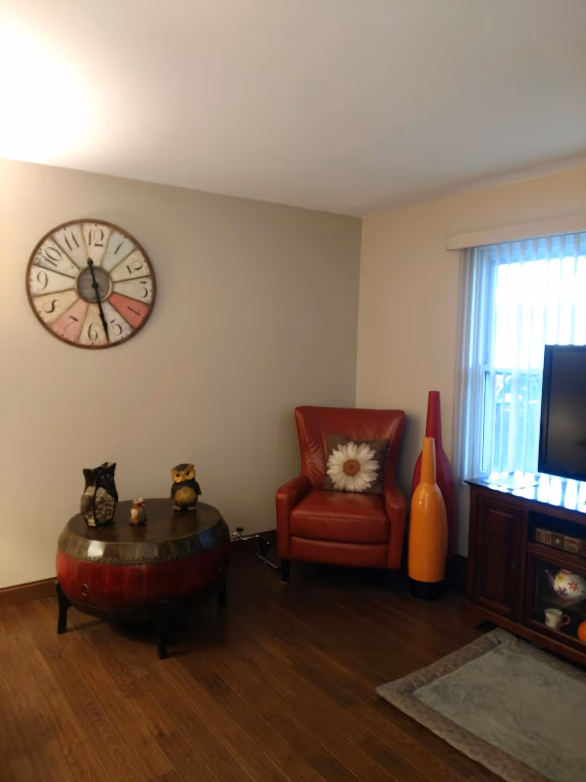 Cozy living room corner with a red armchair, round wooden coffee table with owl figurines, a large wall clock, and a TV cabinet by a window.