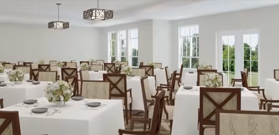 Bright dining room with multiple white-clothed tables, wooden chairs, place settings, and floral centerpieces by large windows.