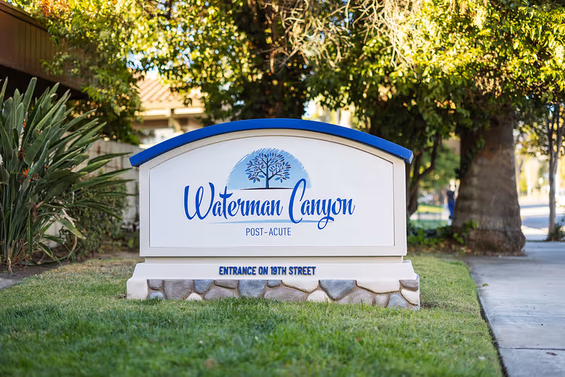 Outdoor sign for Waterman Canyon Post-Acute facility with a blue and white design, situated on a grassy area near a sidewalk and surrounded by trees and plants.